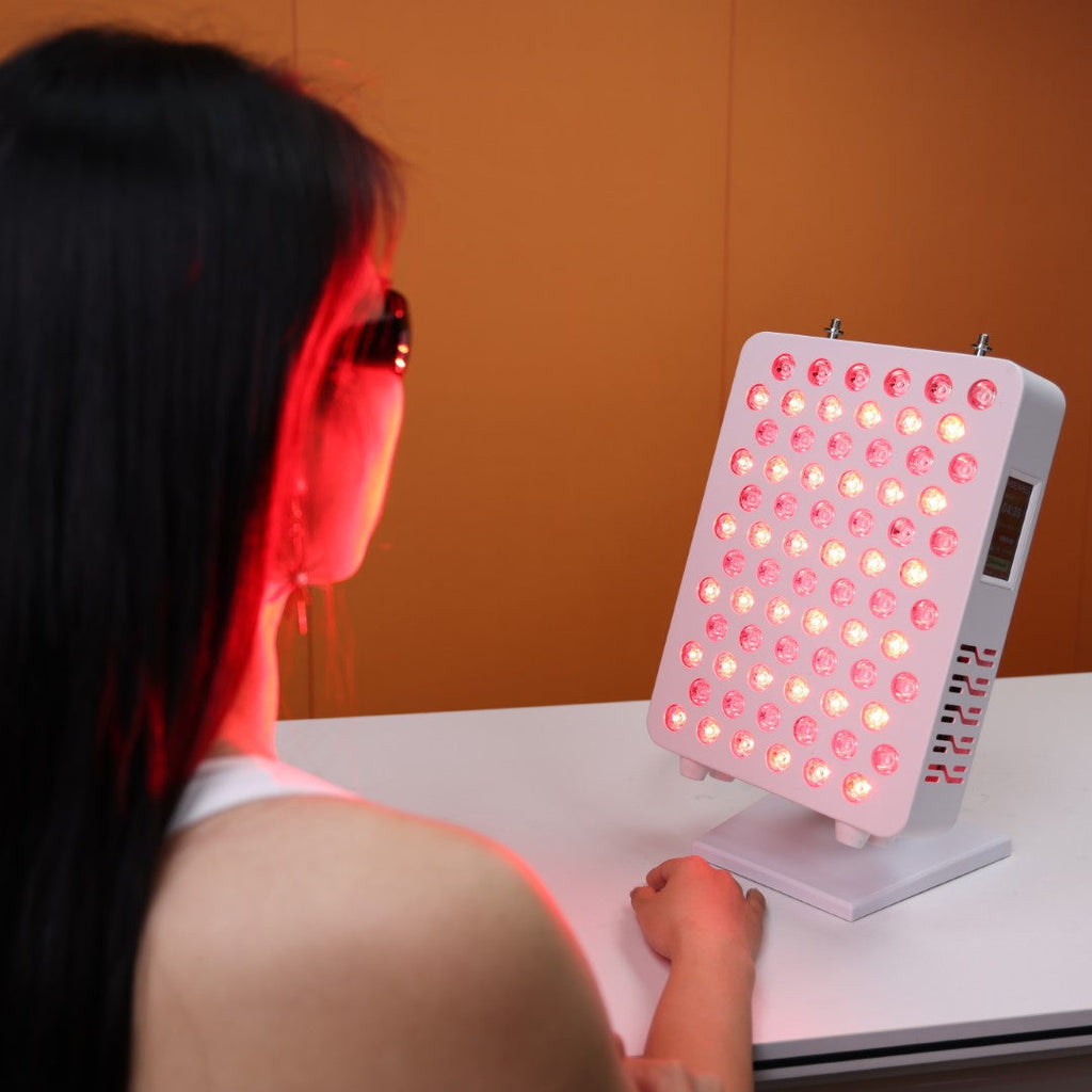 A user engaging in a red light therapy session with the Nicebeam Red Light Therapy Panel. The panel emits therapeutic red and near-infrared light, illuminating the user's face as they wear protective glasses for safety. The sleek panel is positioned on a table with an orange backdrop, showcasing its compact and user-friendly design.