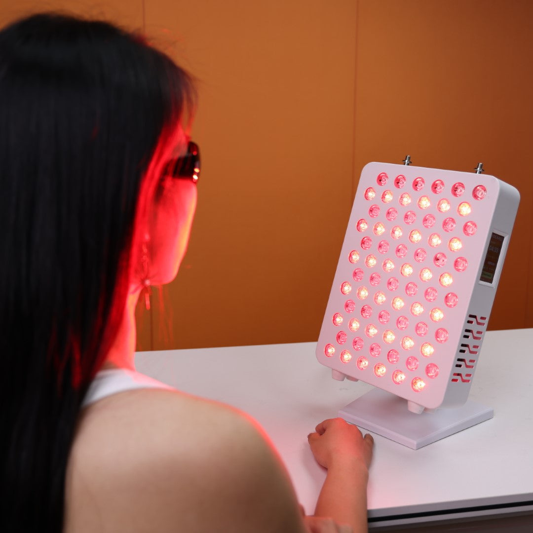 A user engaging in a red light therapy session with the Nicebeam Red Light Therapy Panel. The panel emits therapeutic red and near-infrared light, illuminating the user's face as they wear protective glasses for safety. The sleek panel is positioned on a table with an orange backdrop, showcasing its compact and user-friendly design.